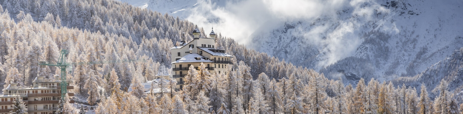 Paesaggio montano innevato con edifici, alberi e una montagna sullo sfondo, cielo blu con alcune nuvole.