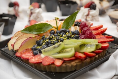 Vibrant fruit tart on a black plate, featuring strawberries, kiwis, and more, with a green leaf, set in a celebratory atmosphere.