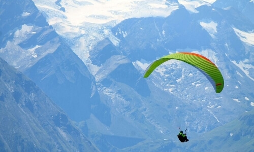 A paraglider with a green and red parasail soars over a stunning mountain range with snow-capped peaks and valleys.