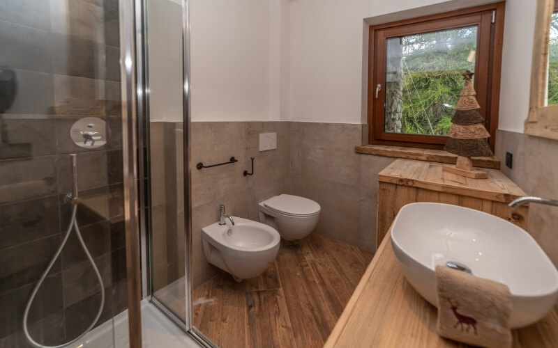 Modern bathroom with white and gray walls, dark wood floor, large window, wooden vanity, shower, toilet, and bidet.