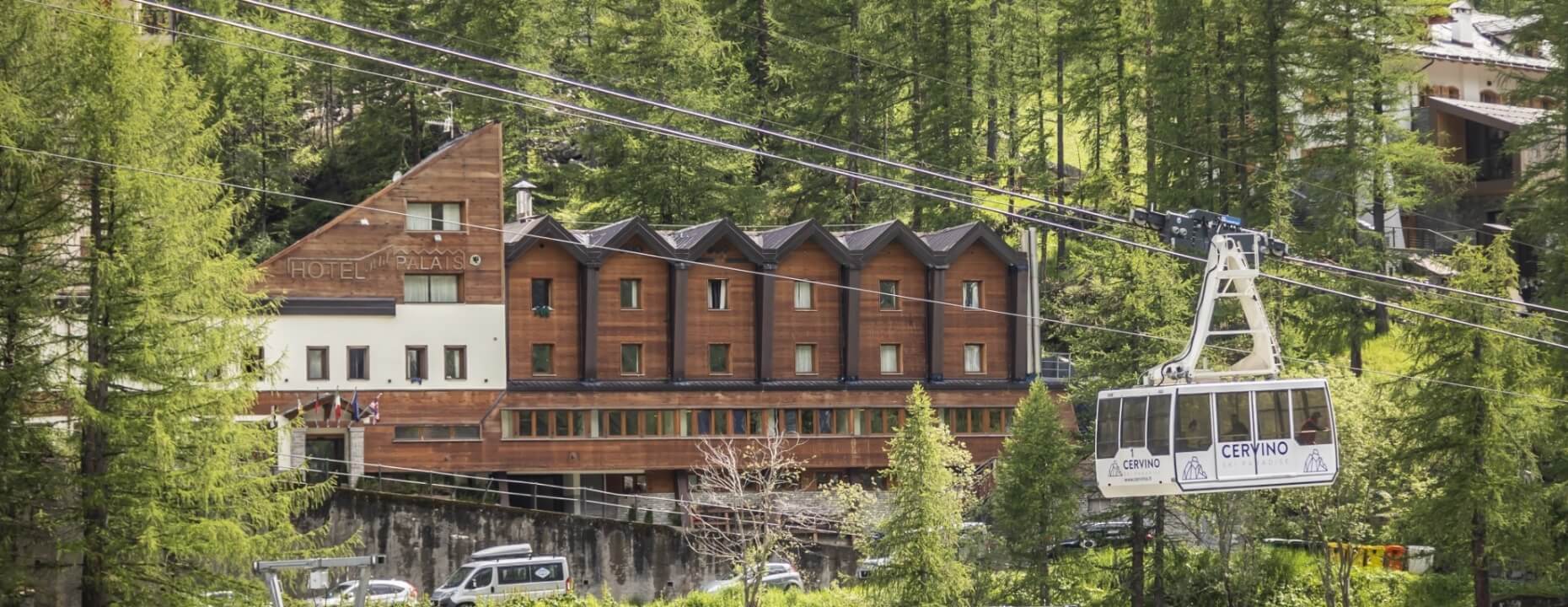 Paesaggio montano sereno con l'Hotel Xalbiso, una funivia e alberi verdi, mentre due furgoni passano sotto.