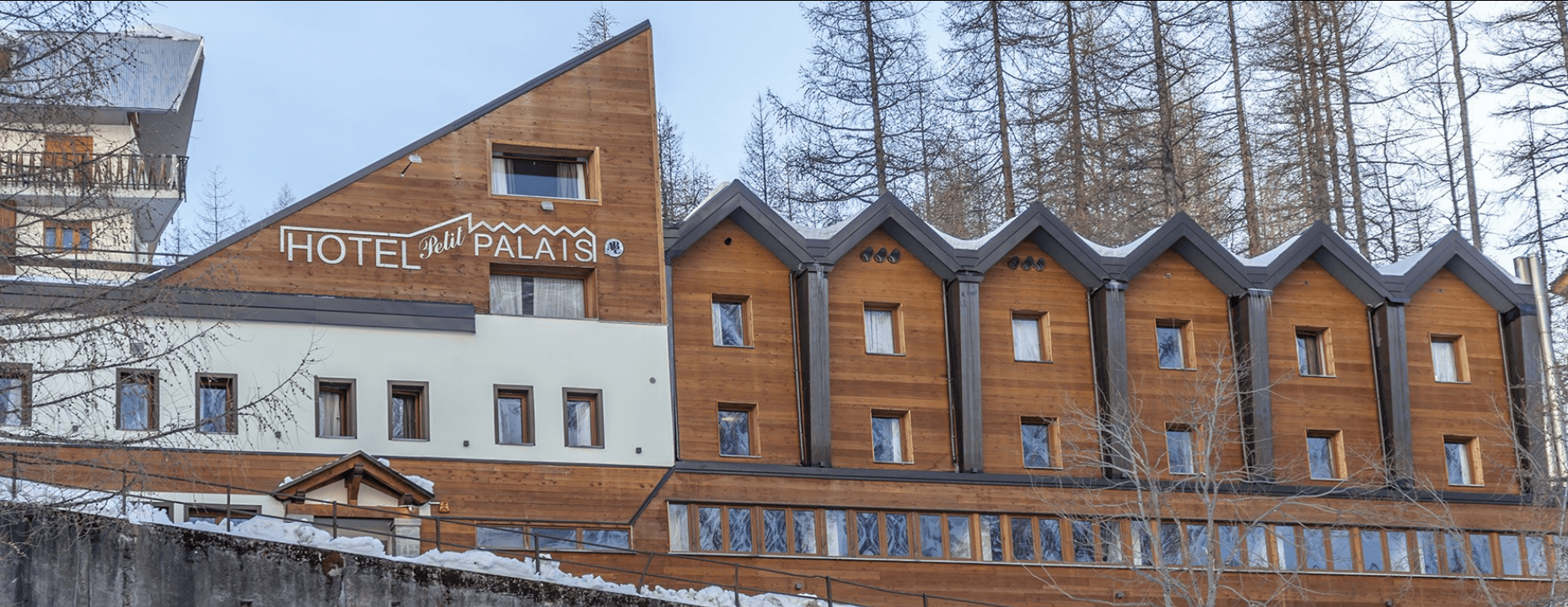 Modern hotel "Petit PALAIS" with wooden exterior, snowy landscape, and clear blue sky, surrounded by tall trees.