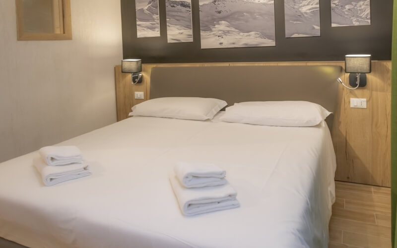 Serene hotel room with a double bed, gray headboard, sepia mountain prints, and light wood tiles, exuding comfort and style.