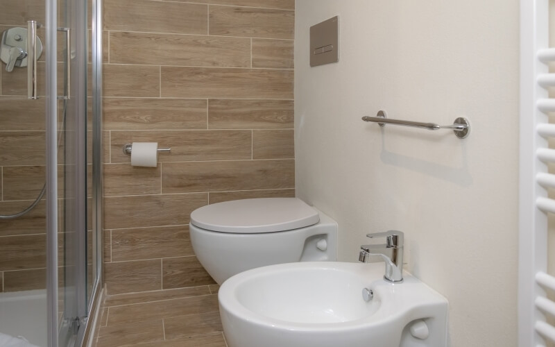 Modern bathroom with shower, toilet, bidet, and towel rack, featuring wood-grain tiles and a cream-colored wall.