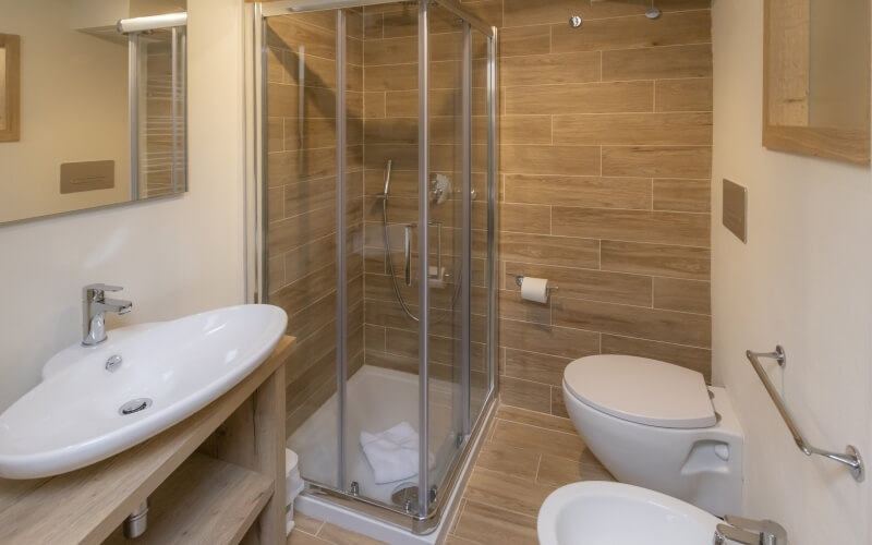 Bagno moderno con doccia in vetro, wc bianco, lavabo su piano in legno e pareti beige. Telo bianco nella doccia.