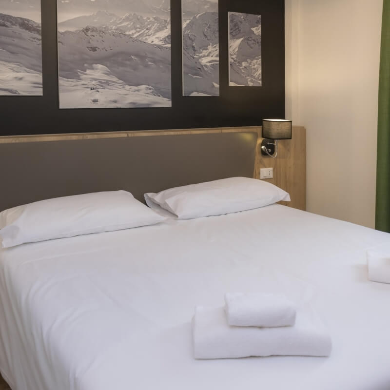 Camera d'hotel serena con letto bianco, cuscini, asciugamani arrotolati e fotografie di montagne sulla parete.
