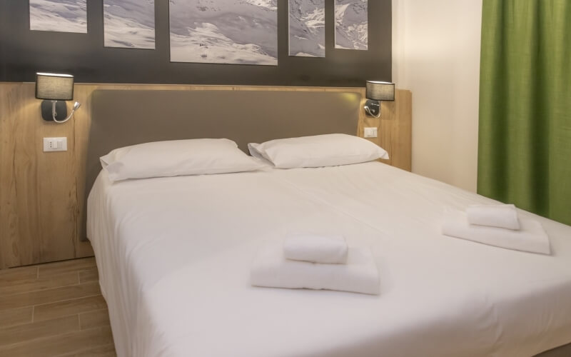 Serene hotel room with a neatly made bed, mountain pictures, wall-mounted lights, and a green curtain against a cream wall.