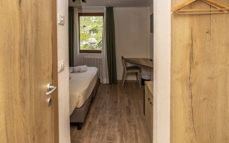 Cozy hotel room with an open door, featuring a bed, desk, and green curtains, illuminated by natural light.