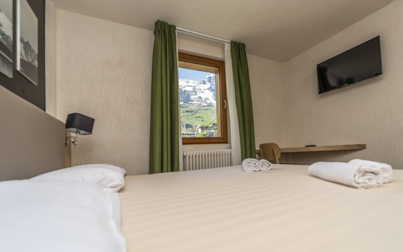 Serene hotel room with a double bed, mountain photo, green curtains, desk, and a view of snow-capped mountains.