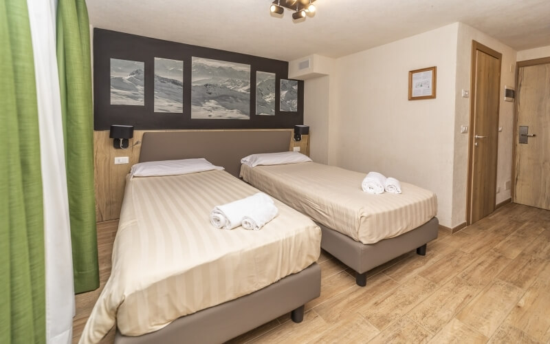 Hotel room with two beds, black and white mountain paintings, wooden door, and green curtain partially covering the view.