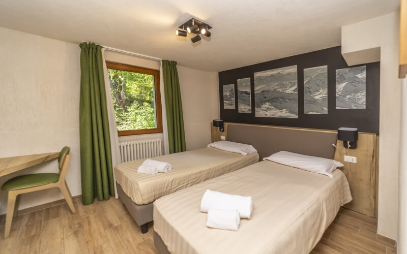 Compact hotel room with two beds, a window, a chair, and mountain paintings on white walls with light wood flooring.