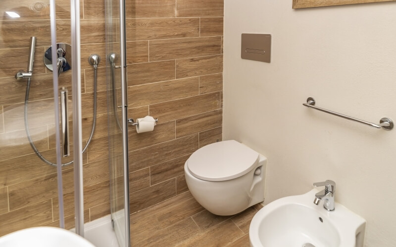 Modern bathroom with glass shower, white toilet, bidet, and light-brown wood-patterned tiles on walls and floor.