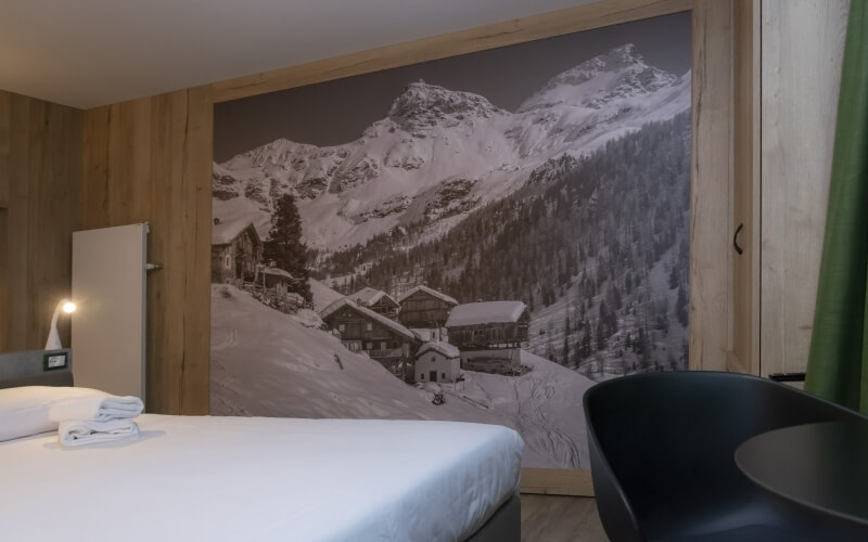 Cozy hotel room with a bed, black chair, snowy mountain mural, warm lamp, and inviting atmosphere for relaxation.