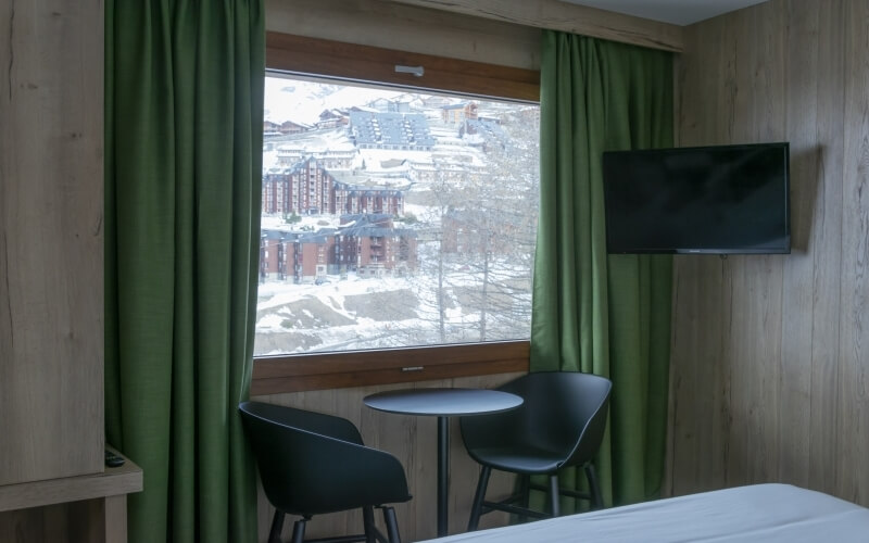 Cozy hotel room with a large window, green curtains, a round table, and a snowy view of surrounding buildings.