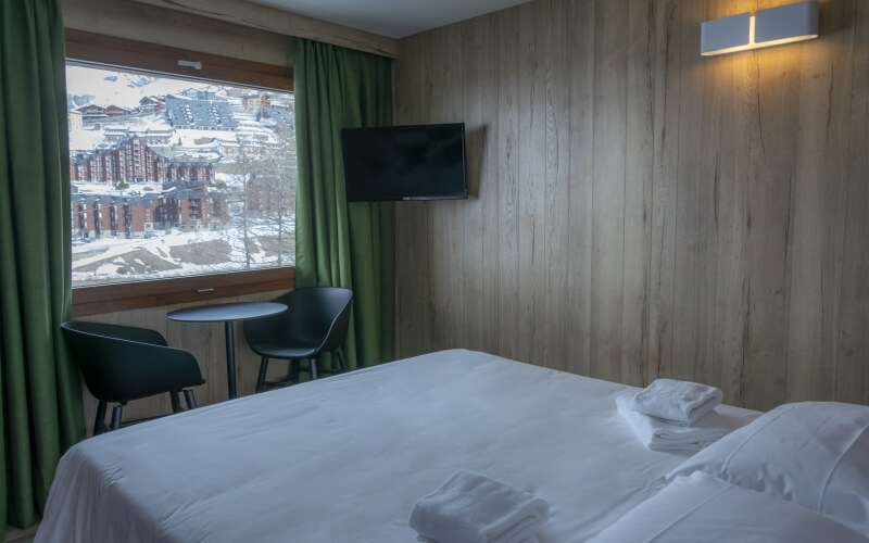 Serene hotel room with a plush bed, minimalist decor, and stunning view of snow-covered mountains through elegant green curtains.