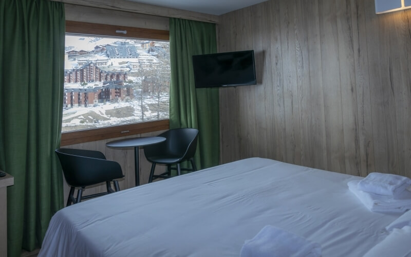 Cozy hotel room with a double bed, flat-screen TV, green curtains, and snowy views, creating a welcoming atmosphere.