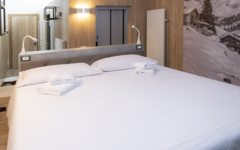 Cozy hotel room with a large bed, lamps, wooden wall, mountain photo, and visible bathroom through an open door.