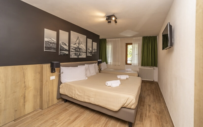 Hotel room with two beds, a window with green curtains, a mounted TV, and mountain paintings on the wall.