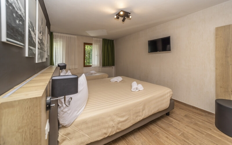 Cozy hotel room with two beds, beige bedspreads, black lamps, a TV, green curtains, and wooden flooring.