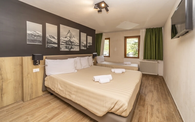 Cozy hotel room with a double bed, single bed, window, TV, and mountain photos, featuring a neutral color scheme.