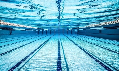 Underwater view of an eight-lane swimming pool with light blue floor, dark blue lane lines, and rippled water surface.