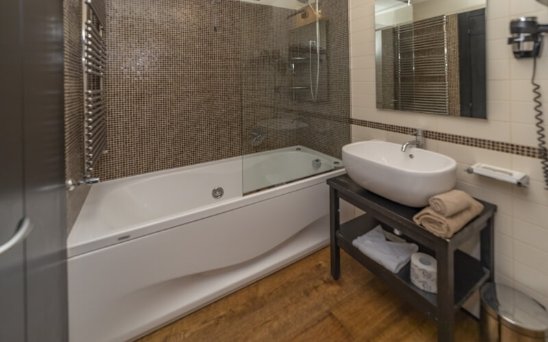 Modern bathroom featuring a white tub, sink with silver faucet, large mirror, beige towels, and light wood flooring.