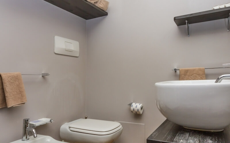 Bagno pulito con WC al centro, lavabo su piano in legno scuro, rubinetti in acciaio e asciugamani appesi.