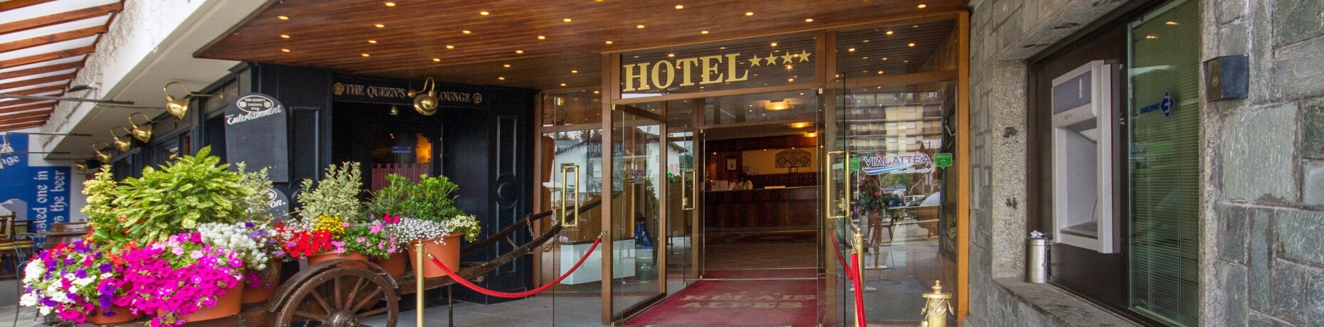 Hotel entrance with glass door, red carpet, stone and wood exterior, colorful flower cart, and welcoming reception visible inside.