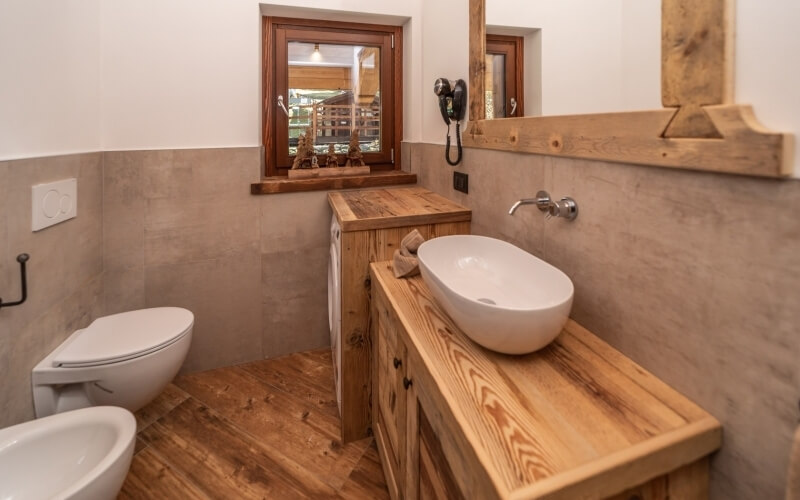 Bagno rustico con toilette bianca, lavabo in legno chiaro, grande specchio e finestra, pavimento in piastrelle marroni.