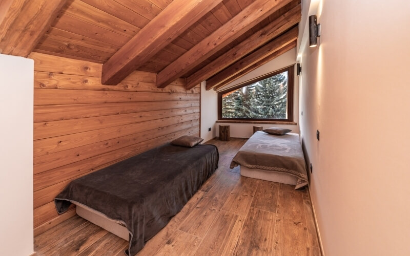 Camera da letto rustica e accogliente con due letti, grande finestra e pareti in legno chiaro, illuminata da luci calde.