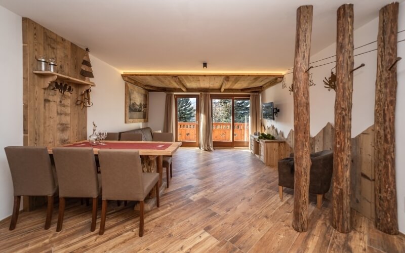 Soggiorno rustico con tavolo in legno, divano marrone, TV a parete e luce naturale che illumina l'ambiente accogliente.