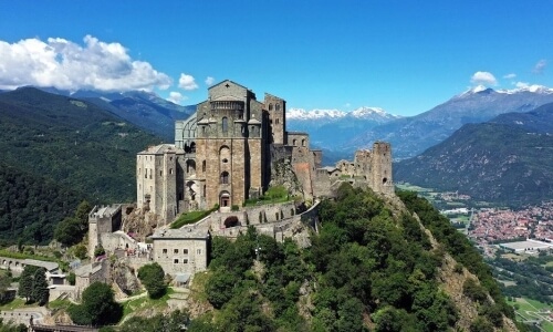 Immagine aerea dell'abbazia Sacra di San Michele nelle Alpi, circondata da montagne e un villaggio in lontananza.