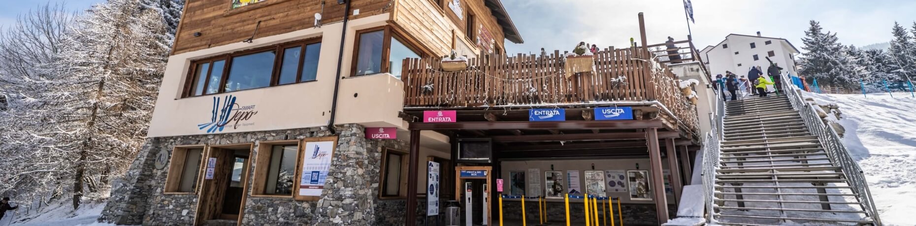 Edificio di un resort sciistico in legno e pietra, con persone sulla scala, alberi innevati e una pista da sci sullo sfondo.