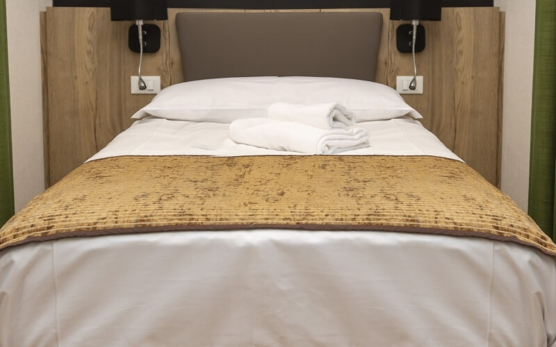 Letto ordinato con lenzuola bianche, cuscini, asciugamani piegati, testiera in legno e coperta beige.