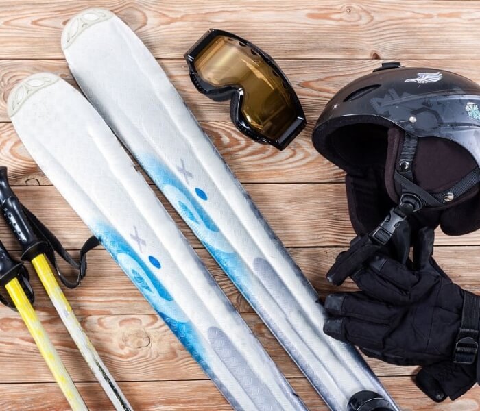 Collection of ski gear: white skis with blue accents, black helmet with visor, black goggles with yellow lenses, and black gloves.