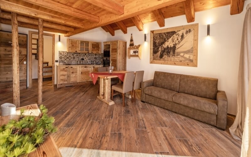 Soggiorno accogliente con cucina, pavimenti e soffitti in legno, divano marrone, tavolo da pranzo e grande quadro sulla parete.