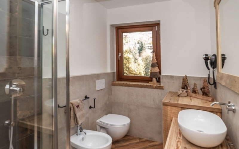 Compact bathroom with two wall-mounted toilets, a wooden vanity, glass shower, and decorative window with pinecones.