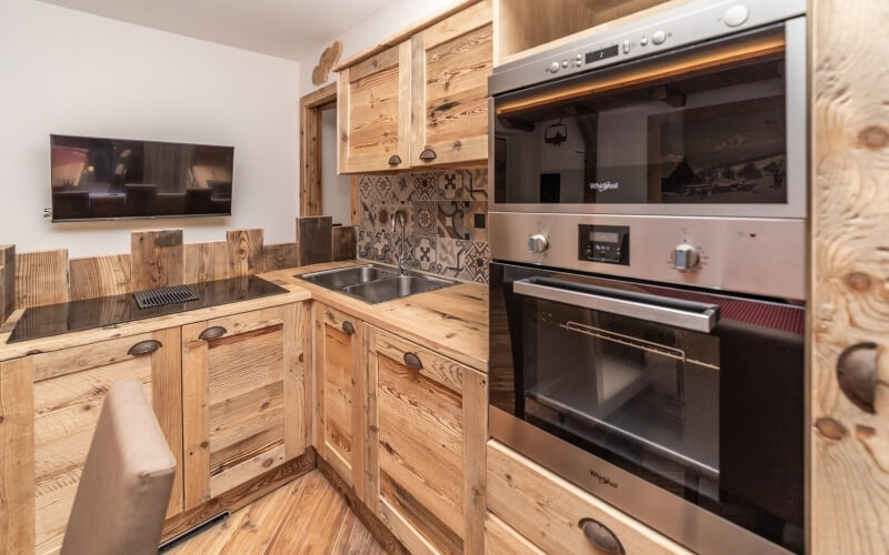 Modern kitchen with wooden cabinets, stainless steel sink, built-in ovens, and a flat-screen TV on a white wall.