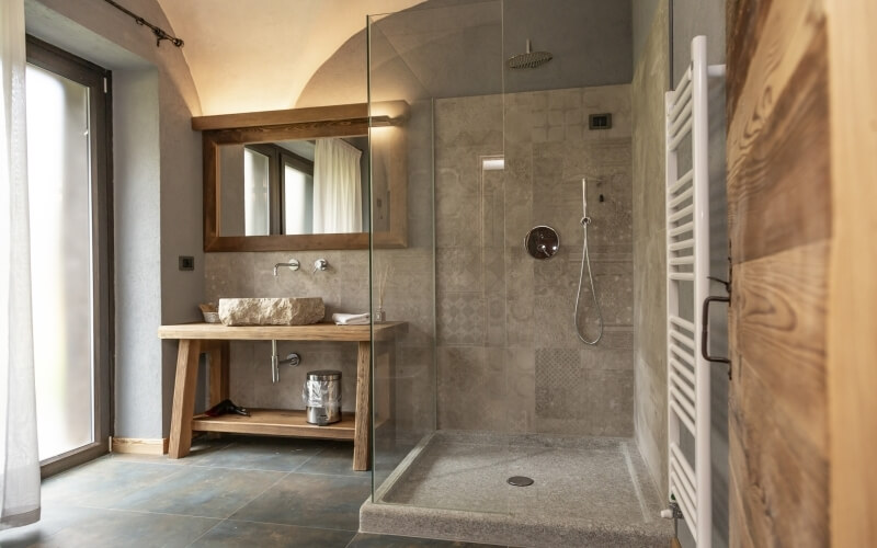 Sleek bathroom with cream vaulted ceiling, wooden vanity, large tiles, and natural light from a window with white curtains.