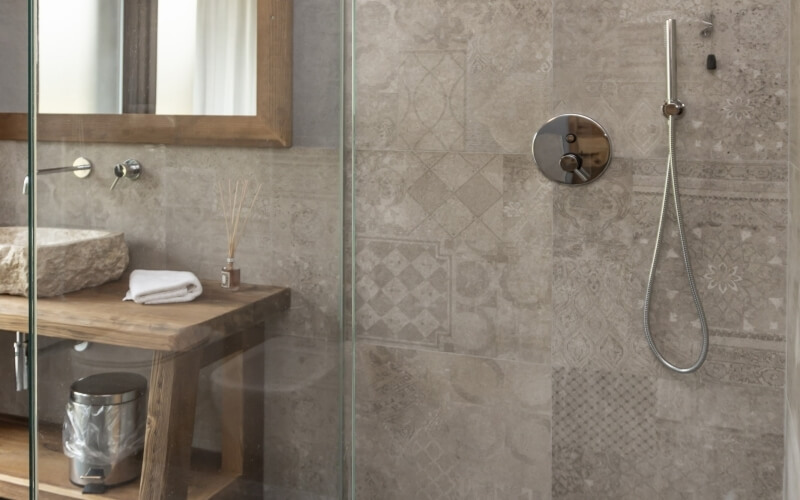 A modern bathroom featuring a stone sink, wooden vanity, mirror, and a patterned shower wall with silver fixtures.