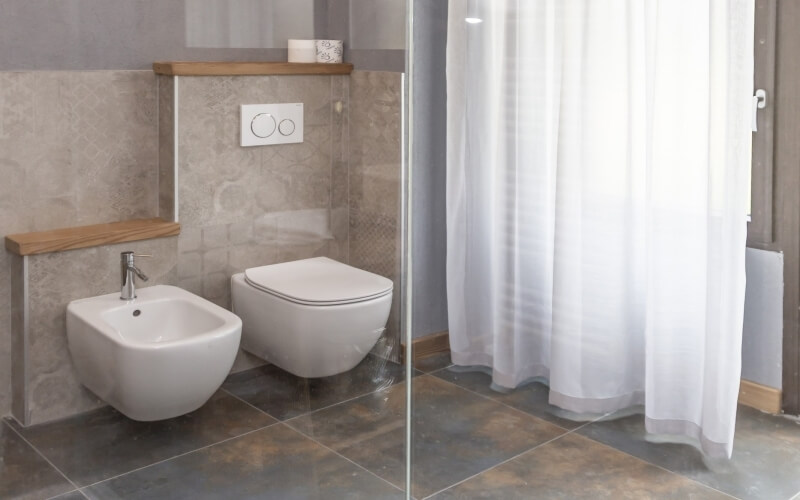 Sectional view of a modern bathroom featuring a glass shower, sleek toilet, bidet, and warm tile accents.