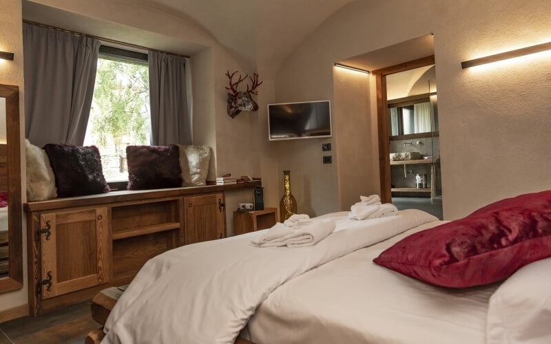 Serene bedroom with gray walls, white linens, red pillows, window seat, and rustic deer head, leading to a darker gray bathroom.