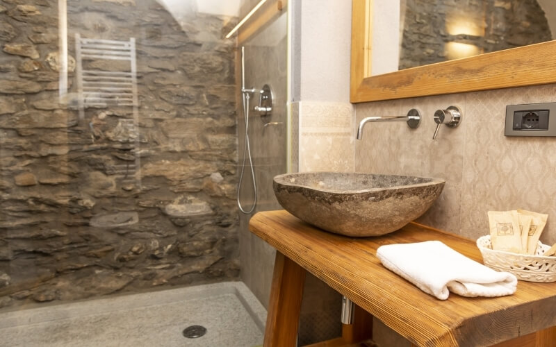 Hotel bathroom featuring a glass shower, stone wall, wooden vanity with a sink, mirror, and toiletries in a basket.