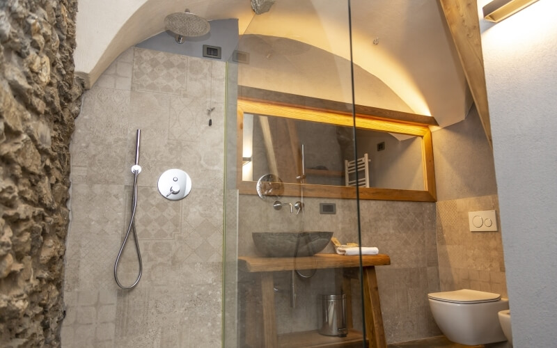 Stylish bathroom in a cave-shaped dwelling with grey tiles, glass shower, wooden vanity, and mixed lighting.