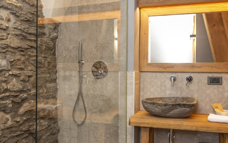 A modern bathroom with a glass shower, stone sink, wooden countertop, mirror, and beige patterned wallpaper.