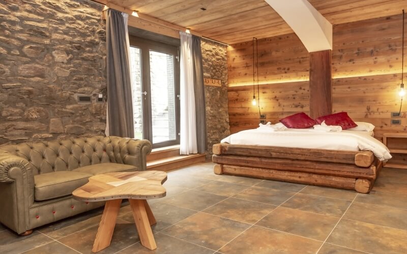 Rustic room with stone walls, wooden accents, a bed with red pillows, tan leather couch, and a wooden coffee table.