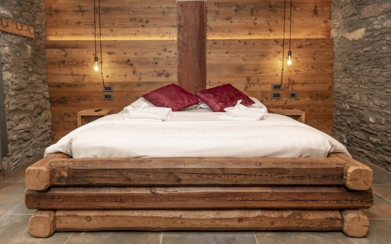 Cozy rustic bedroom featuring a large wooden bed, red pillows, nightstands with lights, and stone walls.