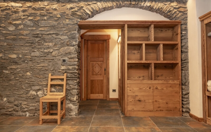 Cozy hallway with stone wall, arched doorway, wooden chair, and built-in bookcase filled with books and objects.