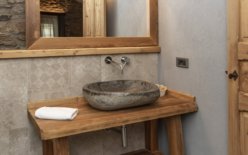 Bagno rustico con lavabo in pietra, accenti in legno, specchio grande e parete di piastrelle grigie. Atmosfera accogliente.