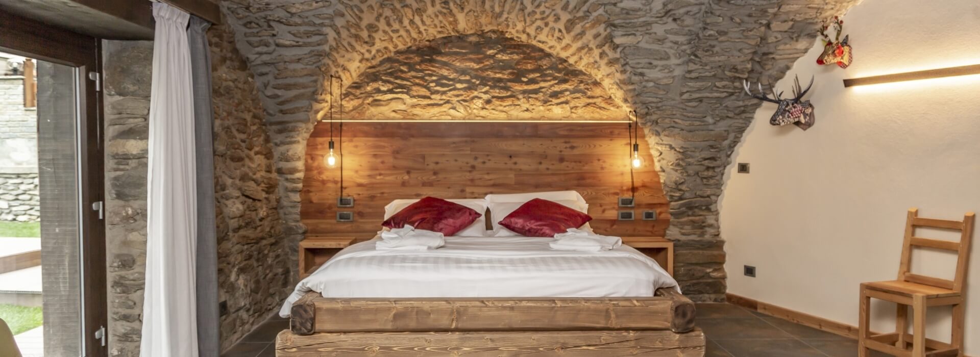 Cozy bedroom with stone wall, wooden bed, white comforter, red pillows, and decorative items, leading to a grassy area outside.